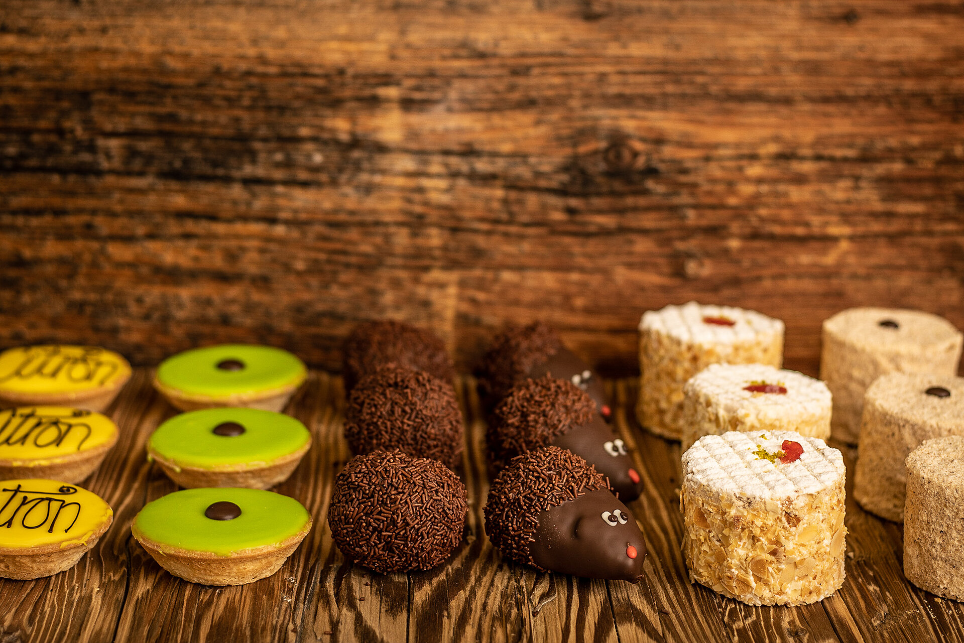 Auf einem dunklen Holzgestell stehen fünf verschiedene Patisserie à je drei Stücke. Links sind gelbe Zitronentörtchen und daneben grüne Caracs. Dann folgen drei Schokoladekugeln mit Streusseln und drei kleine Schokoigel. Es folgen kleine Kirschtörtchen und ganz rechts im Bild sieht man noch Mocca-Törtchen.