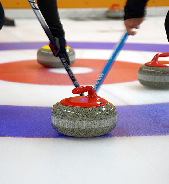 Deux joueurs de curling font glisser une pierre de curling rouge et grise dans un anneau bleu, blanc et rouge. Trois autres pierres de curling se trouvent à côté.