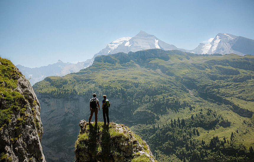 Zwei Wanderer stehen auf einem Felsvorsprung und betrachten die grüne Berglandschaft mit imposanten Gipfeln.