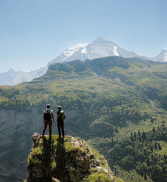 Zwei Wanderer stehen auf einem Felsvorsprung und betrachten die grüne Berglandschaft mit imposanten Gipfeln.