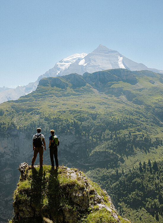 Zwei Wanderer stehen auf einem Felsvorsprung und betrachten die grüne Berglandschaft mit imposanten Gipfeln.