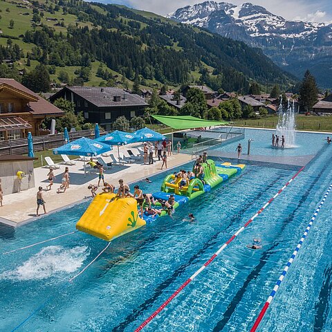 Ein Schwimmbad mit bunten Wasserattraktionen und Menschen, die Spaß haben. Im Hintergrund sind Berge und eine schöne Landschaft zu sehen.